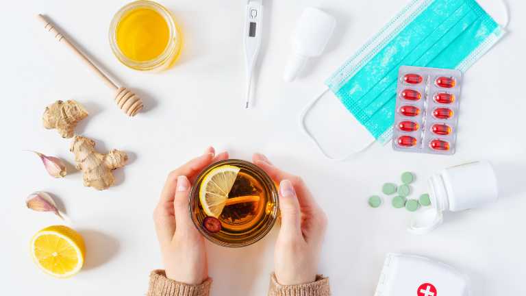A collection of cold and flu treatments, including over-the-counter medicines like cough syrup and tablets, alongside natural remedies such as a steaming cup of ginger and honey tea and a jar of honey with lemon slices.